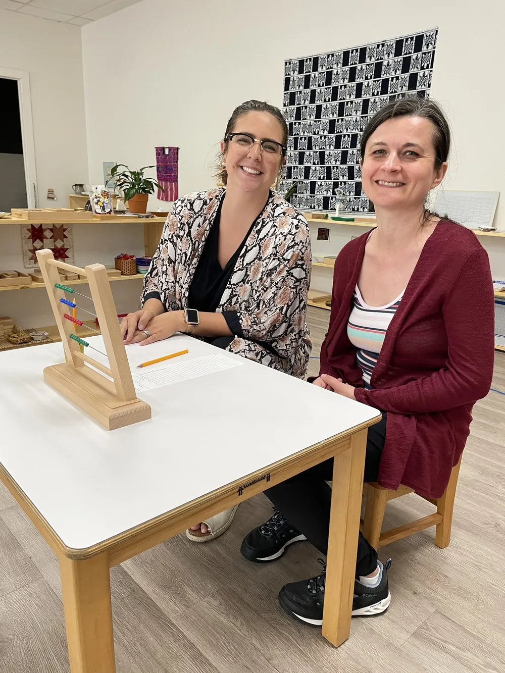 Two smiling adults at a classroom table with a bead frame and a written lesson.