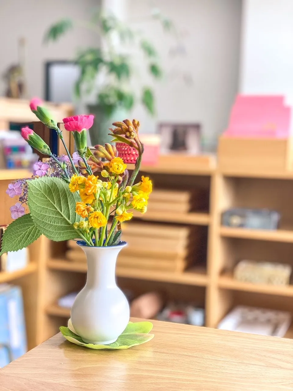 A white vase of pink, yellow, and purple flowers on a wooden classroom table.