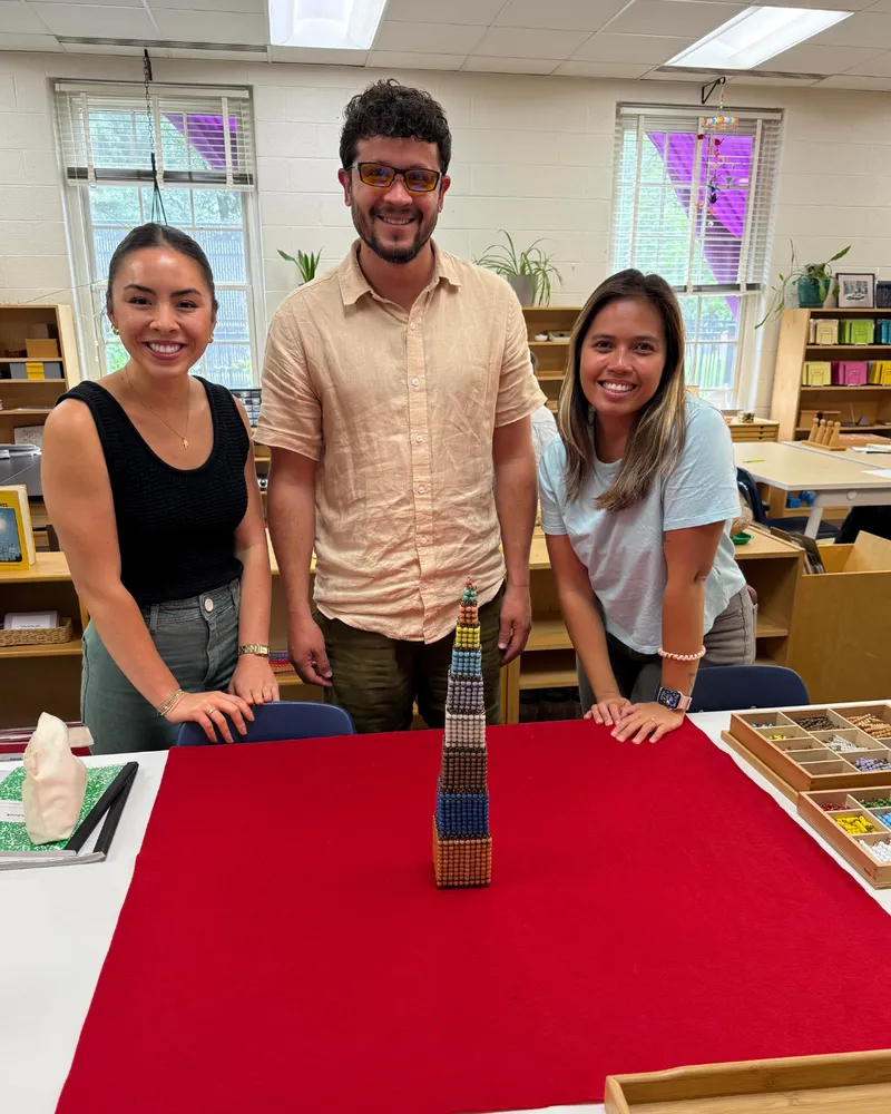 Three smiling adults behind a red work mat holding a stacked tower of Montessori bead cubes.