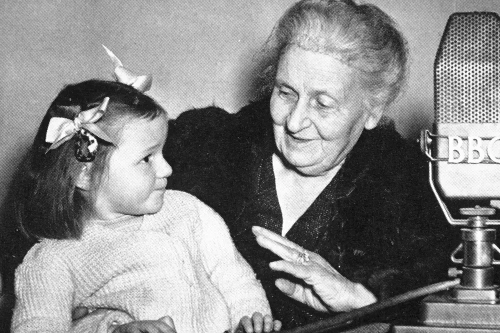 Maria Montessori smiling at a young girl on her lap beside a BBC microphone.
