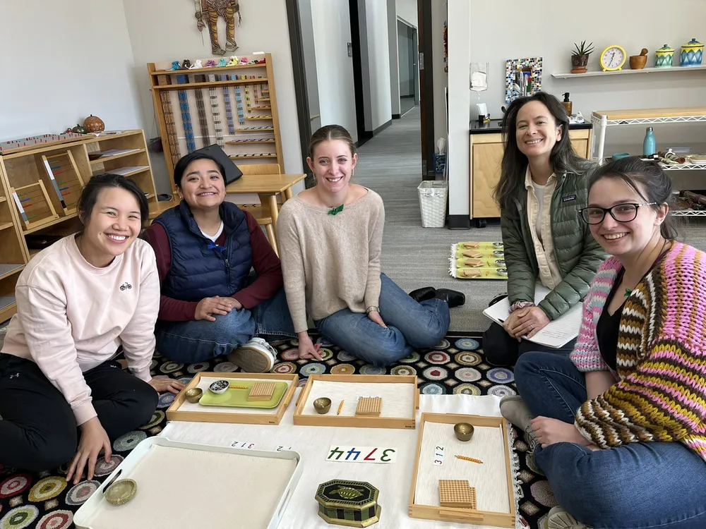 Five smiling adults sitting on a classroom rug with Montessori math trays and numeral cards.