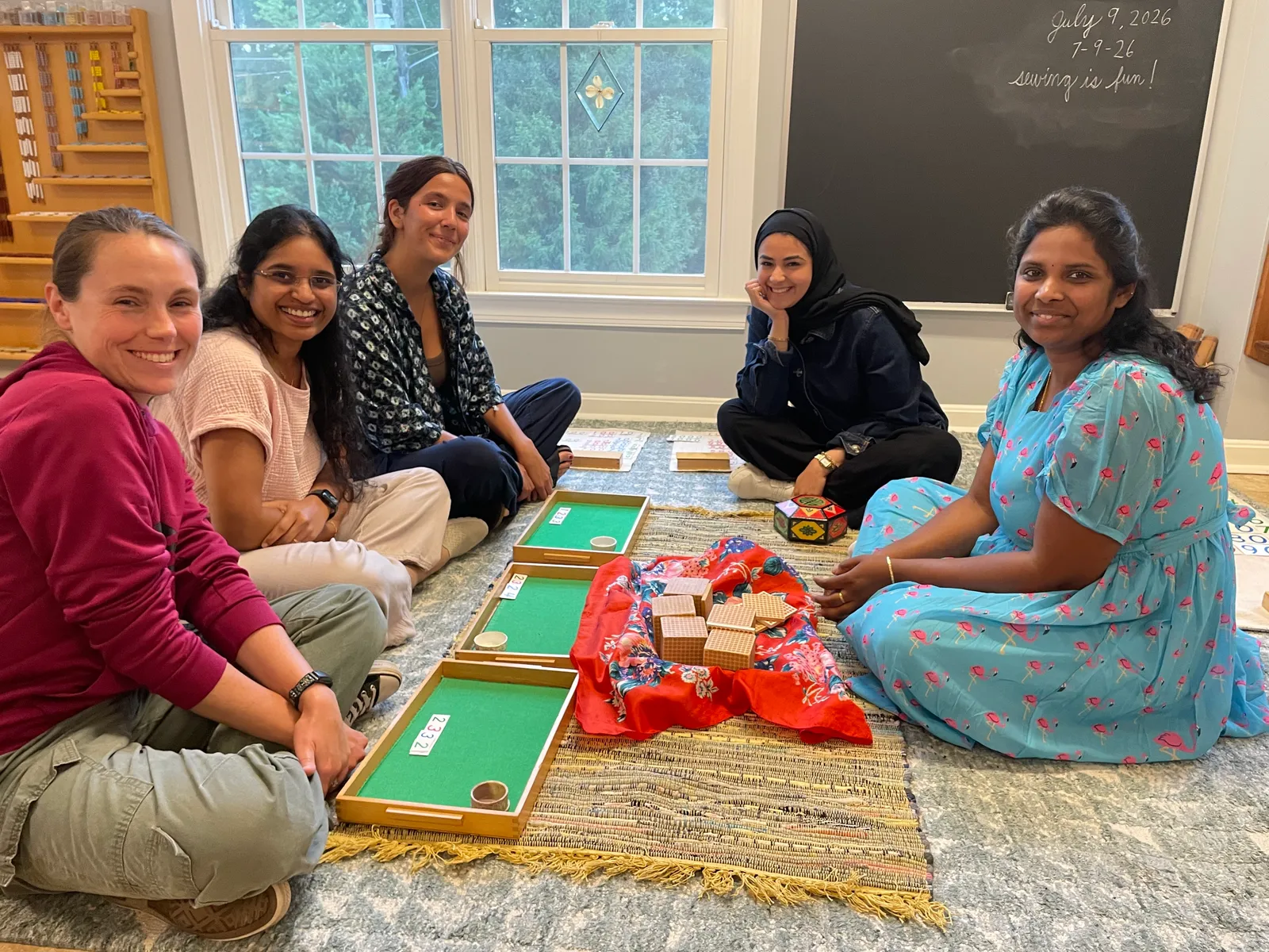Five smiling adults sitting on a classroom rug with green trays and wooden cubes, in front of a chalkboard.