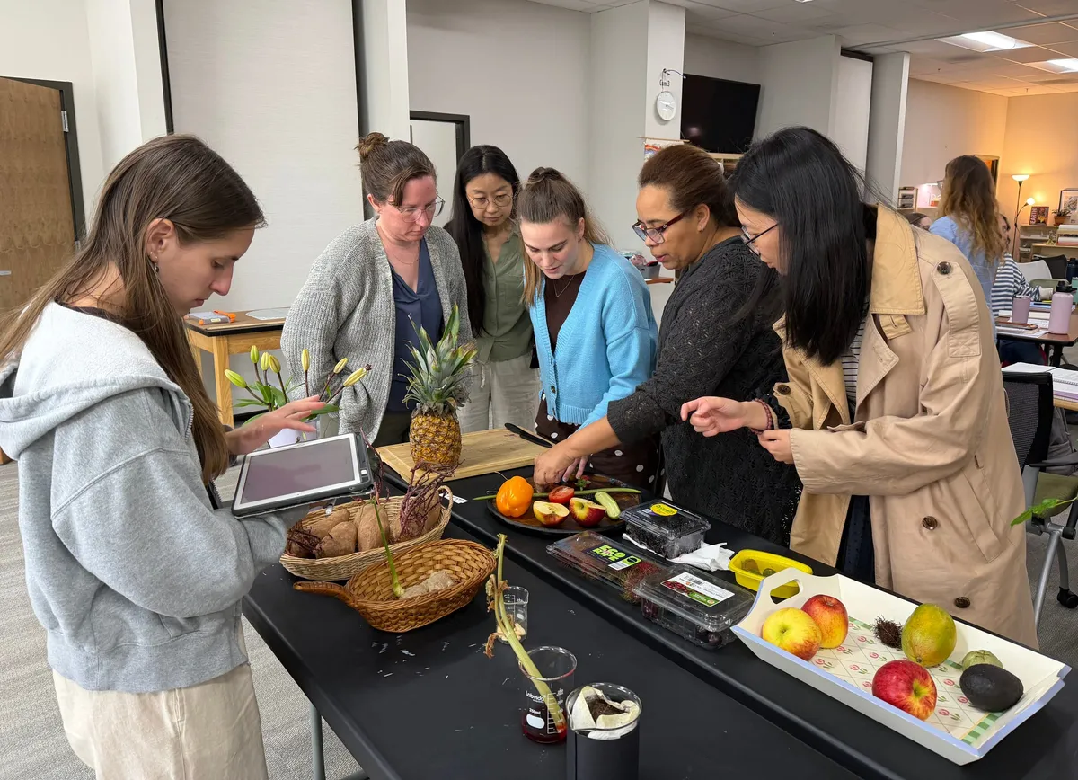 Six adults around a table of fruit, vegetables, and a cutting board, practicing a food preparation activity.